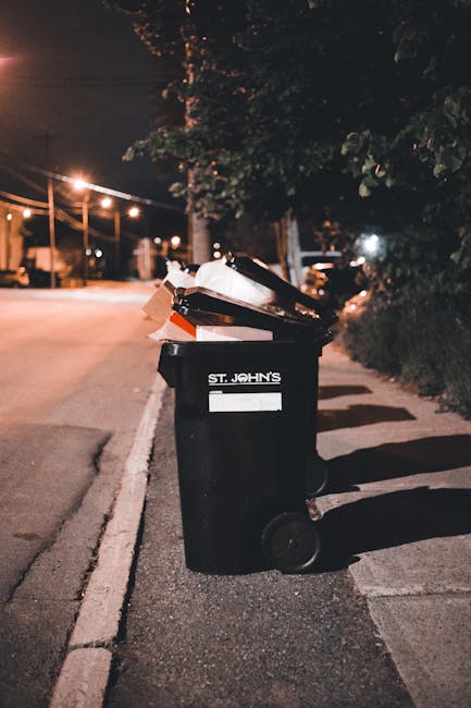 A black wheeled rubbish bin positioned on the pavement beside a street at night, with the label 'St. John's' visible on its side. The bin is open, revealing a mix of various waste items, including car