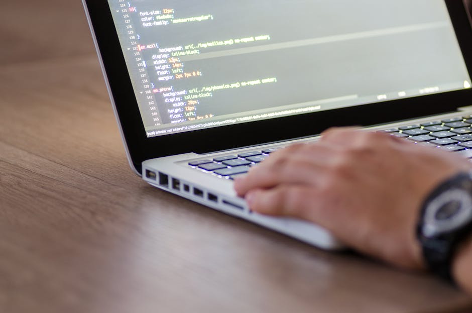 A person seated at a desk working on a laptop, which displays lines of colorful computer code on its screen. The individual is using a mouse with their right hand and has their left hand on the keyboa